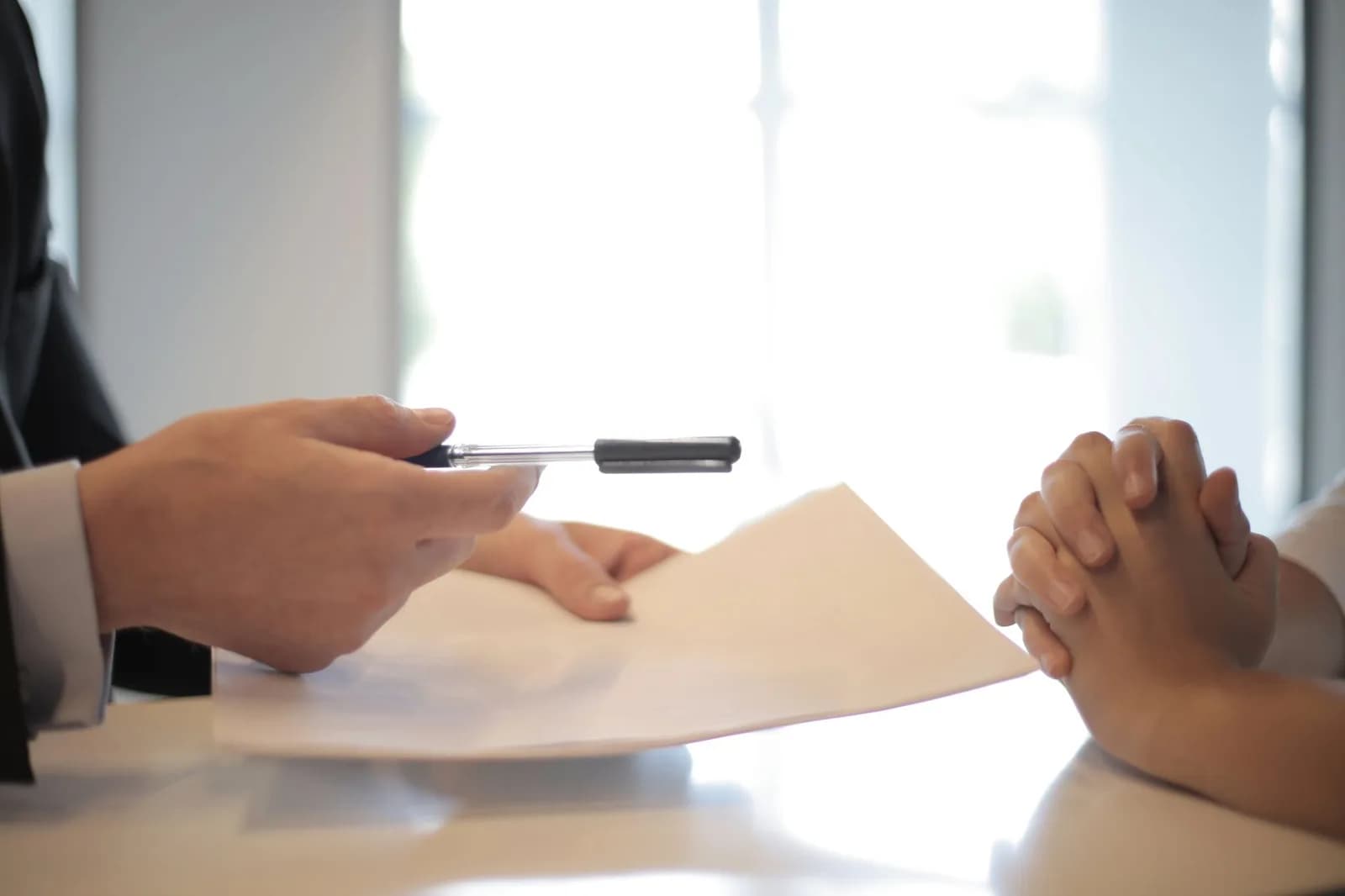 Mão entregando caneta sobre contrato em branco, com mãos entrelaçadas do outro lado da mesa