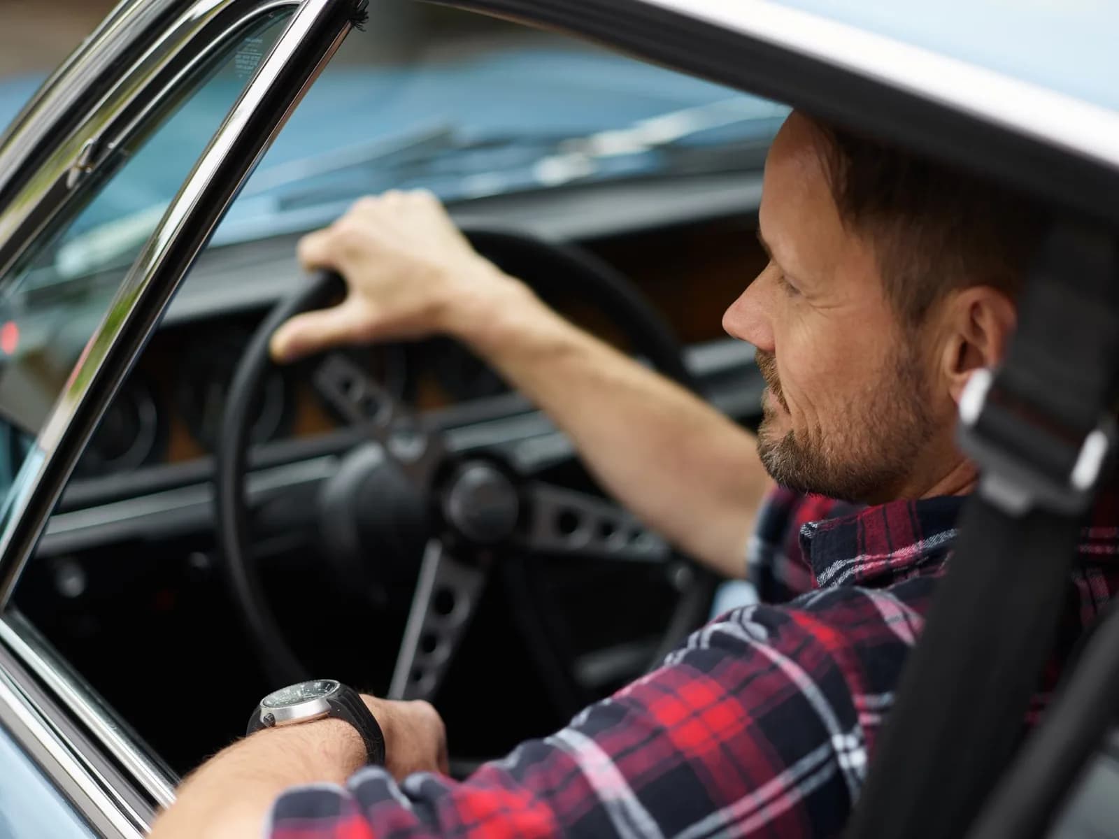 Homem adulto dirige um carro, com as mãos no volante e cinto de segurança visível