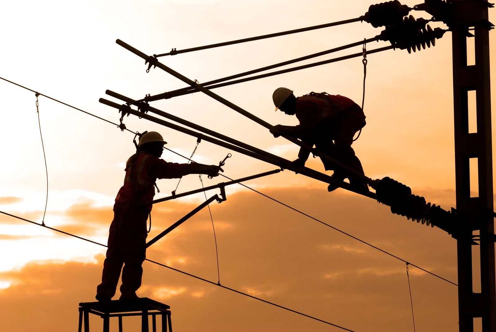 Eletricistas trabalham em rede elétrica ao entardecer, silhueta contra o céu alaranjado