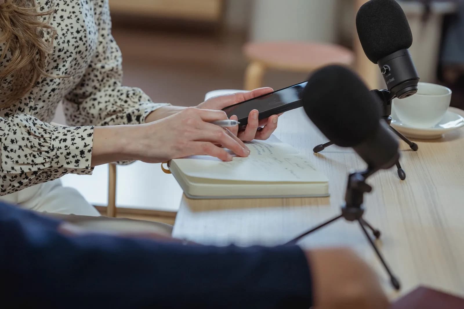 Mão segura o celular gravando, com microfone e caderno de anotações à mesa
