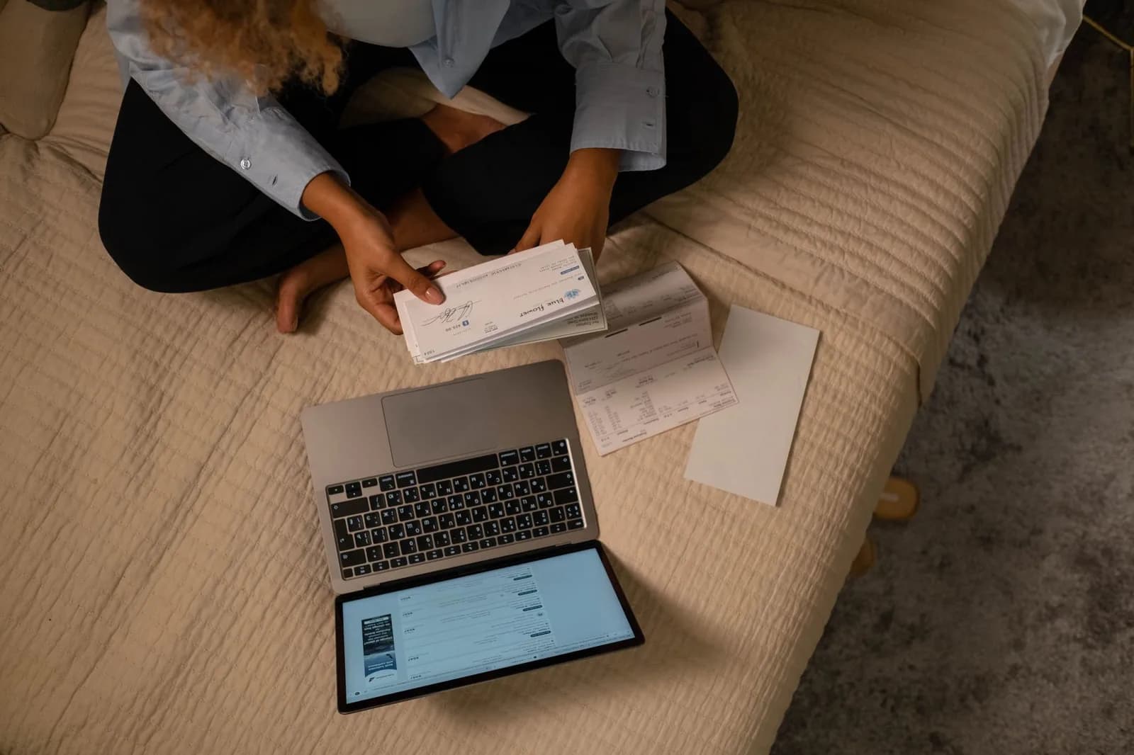 Pessoa sentada com contas e notebook sobre a cama