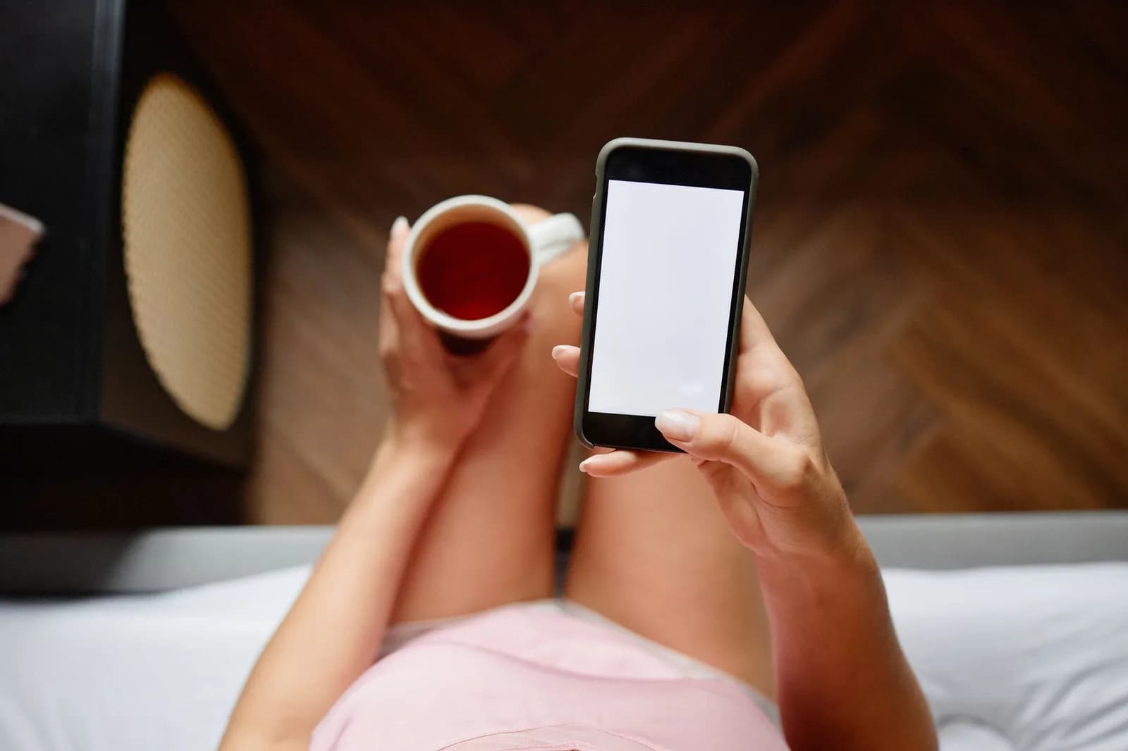 Mão segura smartphone de tela branca, com xícara de chá na outra mão