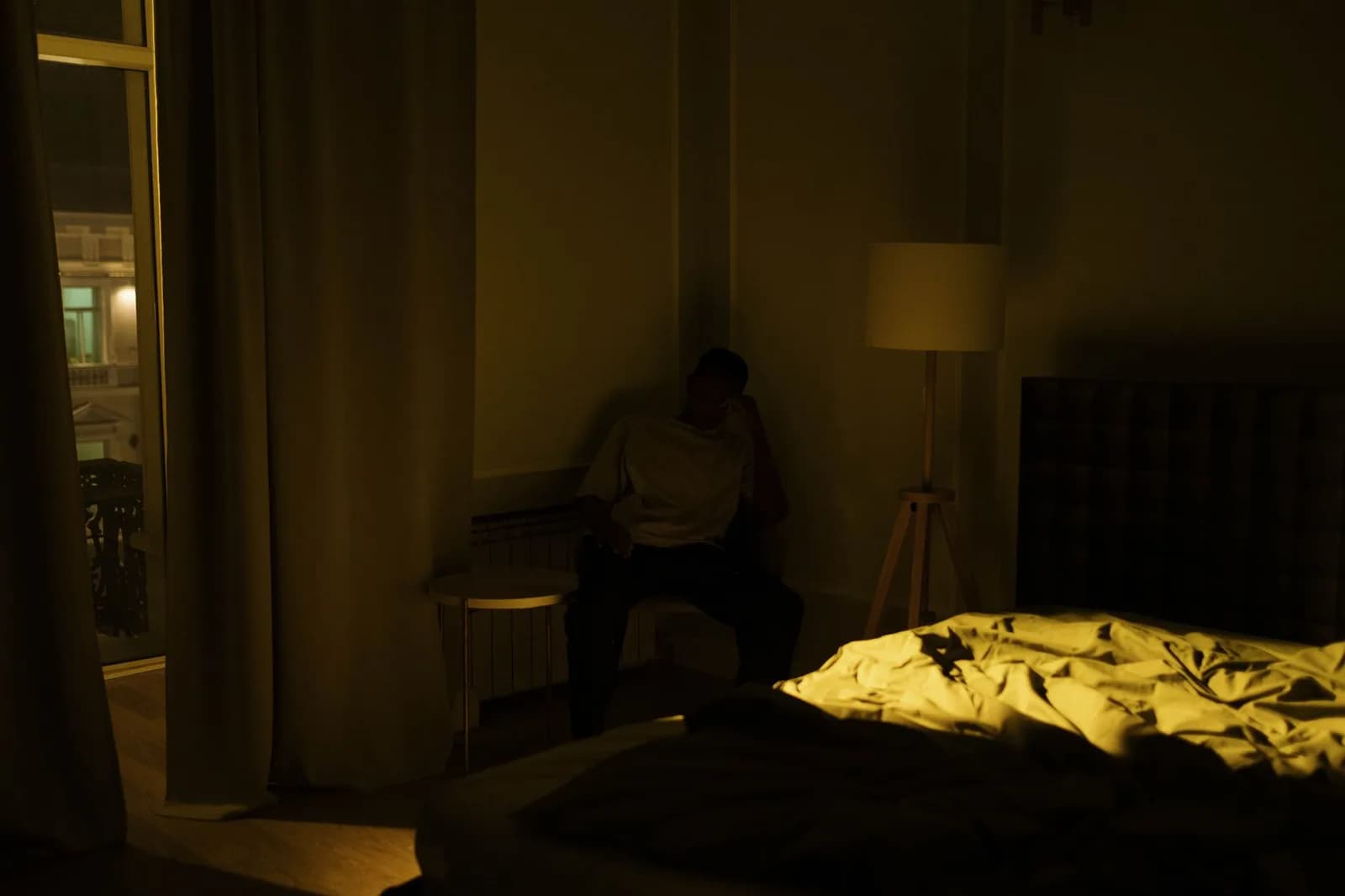 Homem sentado sozinho na beira da cama em quarto escuro, iluminado por um abajur