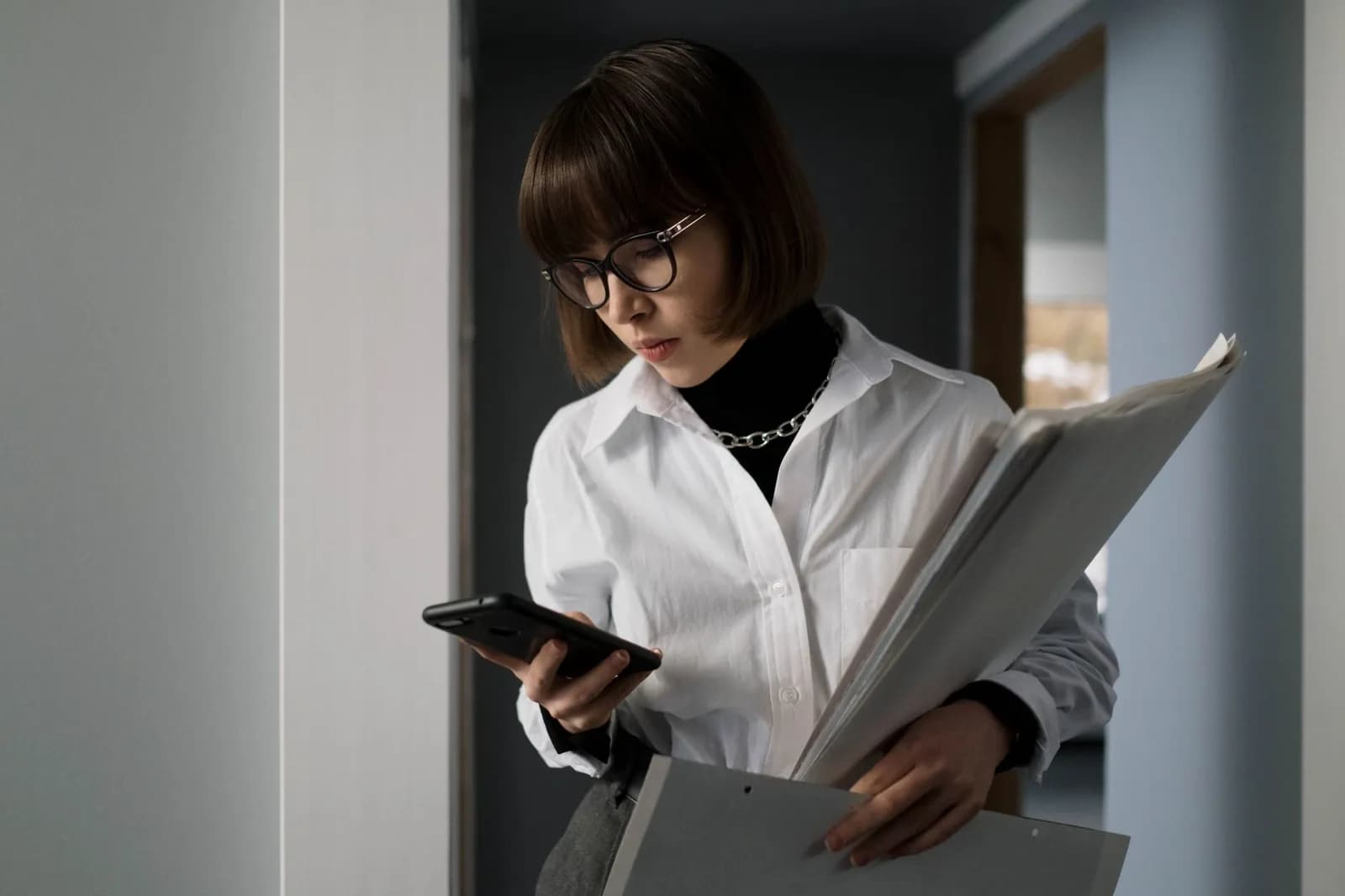 Mulher com pasta de documentos confere o celular em corredor