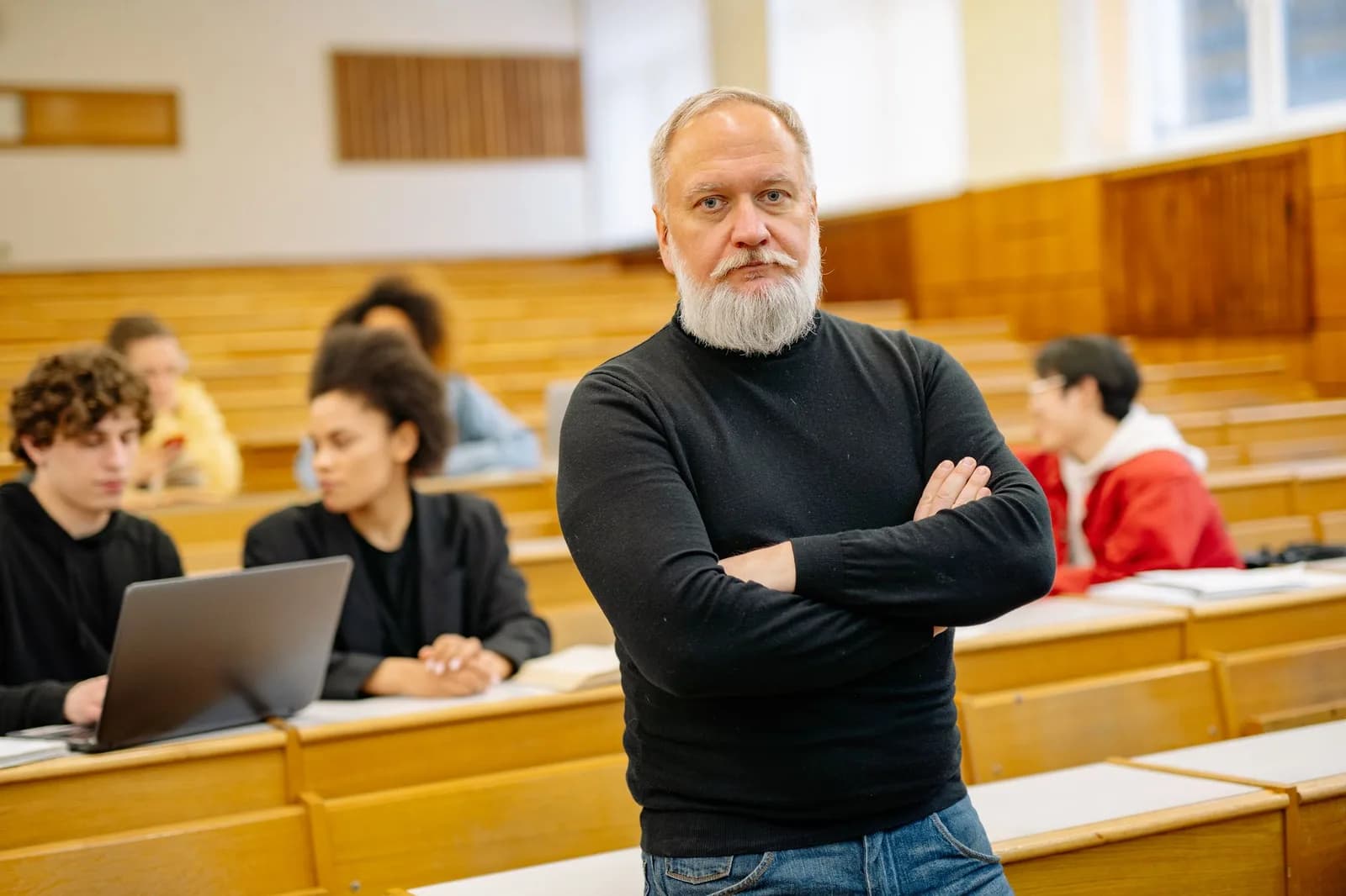 Professor de braços cruzados em um auditório, com estudantes sentados ao fundo