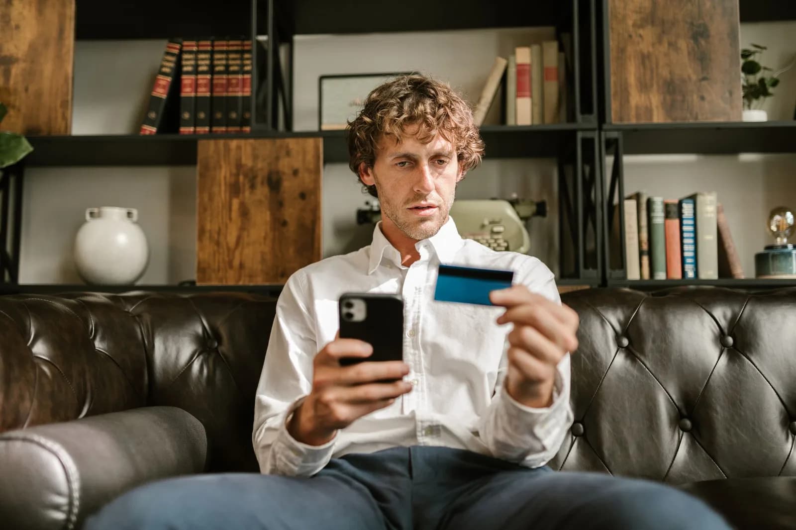 Homem sentado em um sofá de couro segura um celular e um cartão de crédito